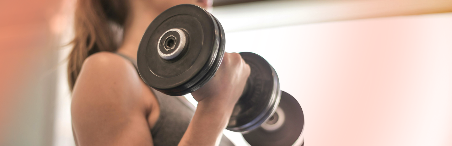 A close-up of a woman lifting a black dumbbell, focusing on her hand and the weight, against a softly blurred background.