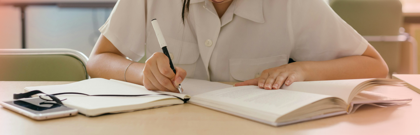 A student in a shirt writes notes in a notebook beside an open textbook and a smartphone on a wooden desk.
