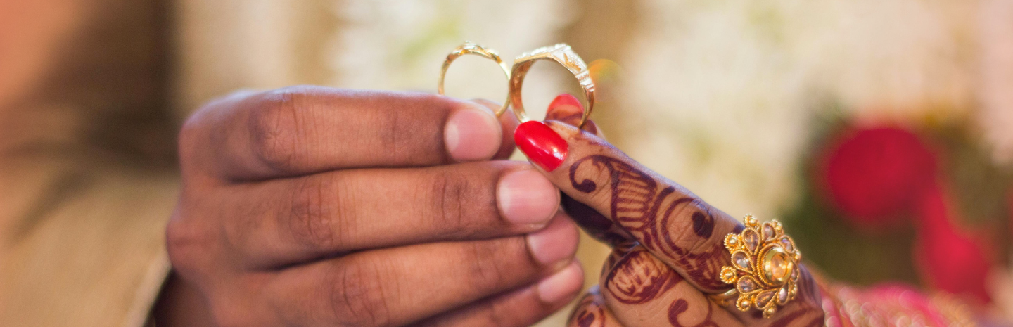 The image shows a close-up of a traditional Indian wedding moment, where a person is placing a ring on another's decorated hand with henna, jewelry, and colorful bangles.