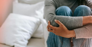 A person sitting on a bed, hugging their knees to their chest.