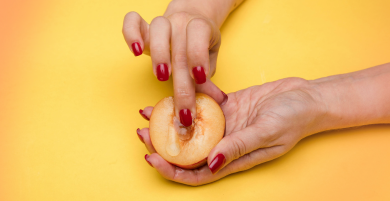 A person with red nail polish presses a finger into the center of a halved peach, suggestive of sexual innuendo, against a yellow background.