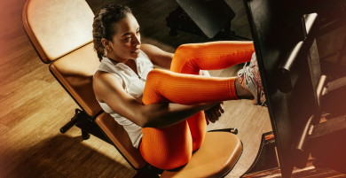 Woman in orange leggings and white top exercises on a leg press machine at the gym. She looks focused and determined, with warm lighting and wood flooring.