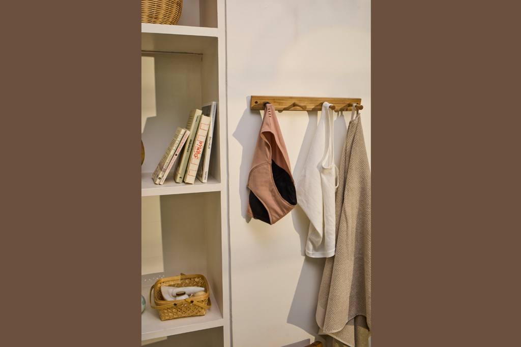 Beige and black period panty hanging on a wooden wall hook alongside white and beige garments, next to a bookshelf with neatly arranged books.