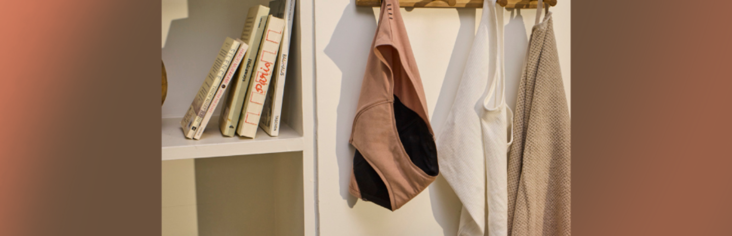 Person holding a beige period panty with black inner lining, surrounded by light-colored fabric and hanging laundry.