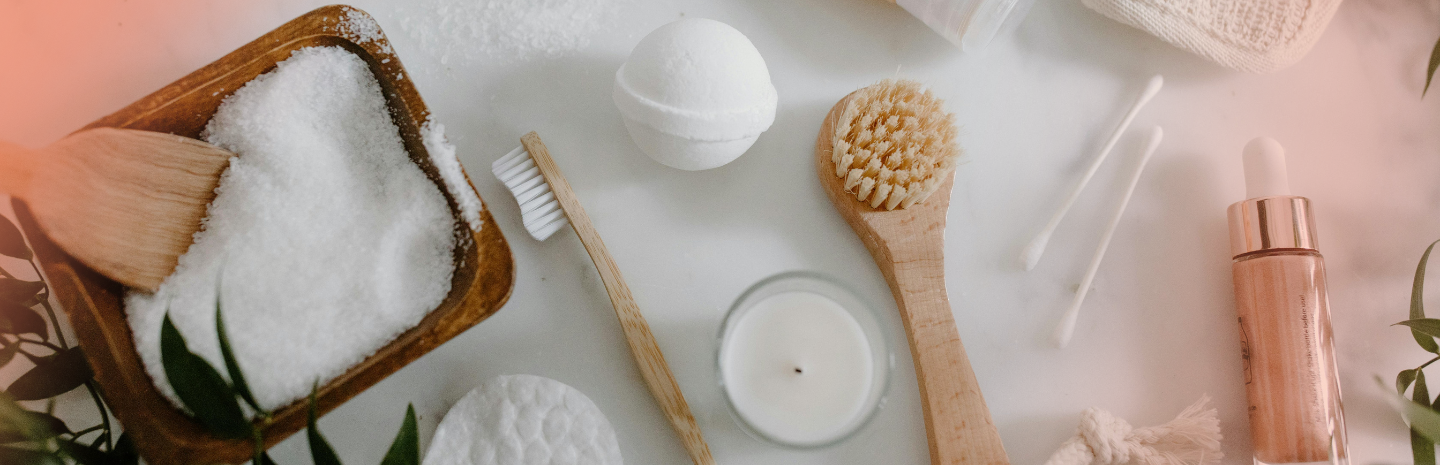 A flat lay of spa products including bath salts, a bath bomb, toothbrush, skincare bottle, and natural items on a marble surface.