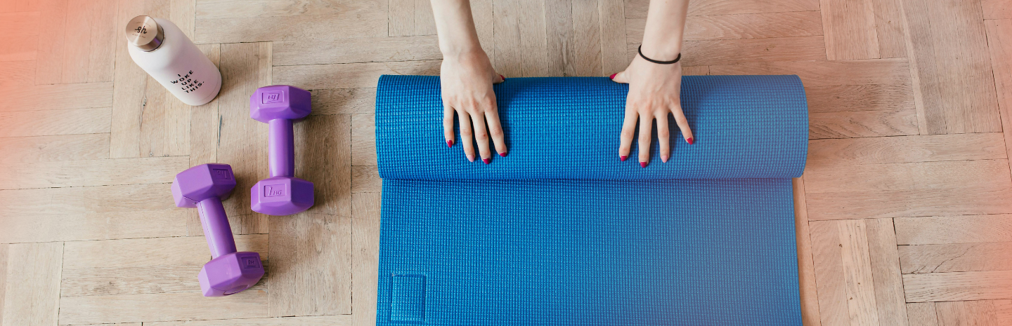A person is rolling up a blue yoga mat on wooden flooring, with purple dumbbells and a water bottle nearby.