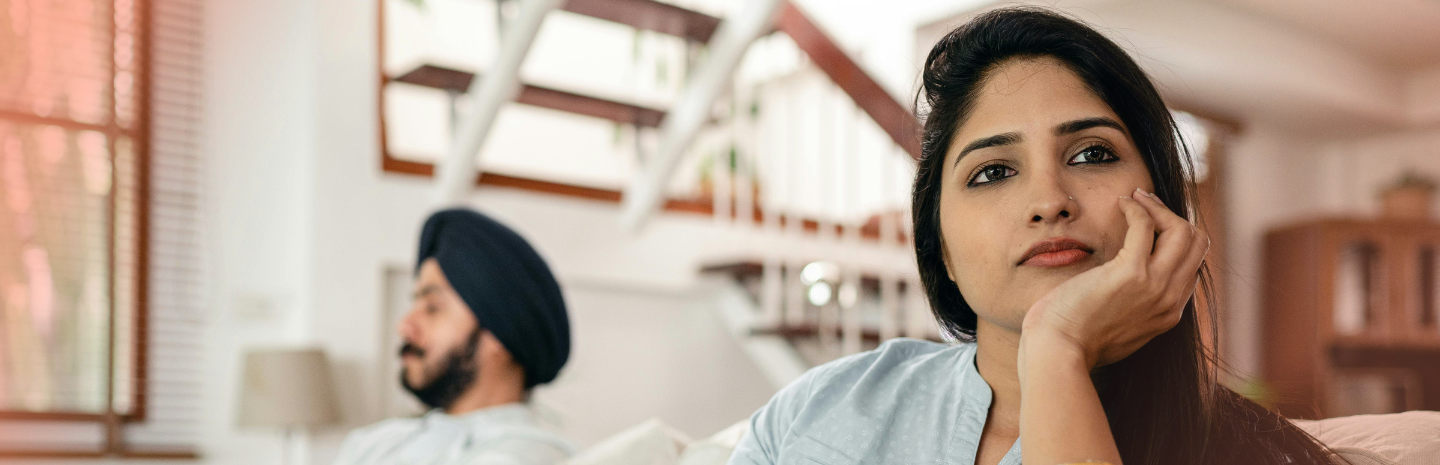 Woman looking upset while sitting on a couch with her partner in the background after an argument