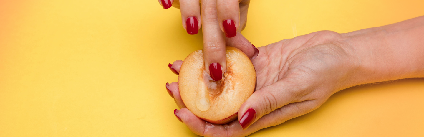 A person with red nail polish presses a finger into the center of a halved peach, suggestive of sexual innuendo, against a yellow background.
