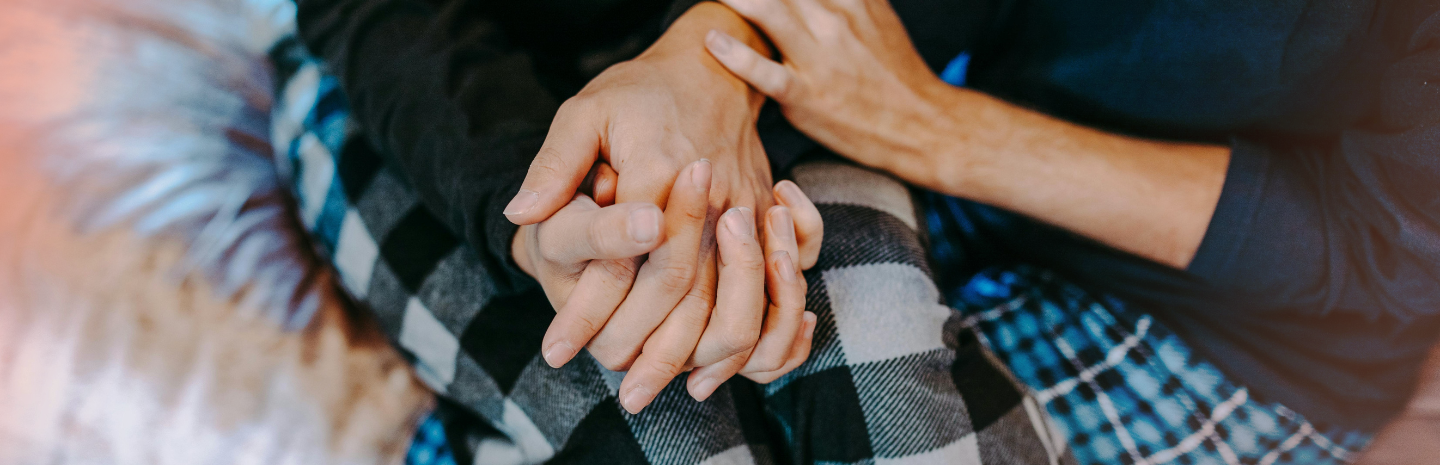 Two people sit close together in plaid clothing, holding hands in a gesture of comfort or support.
