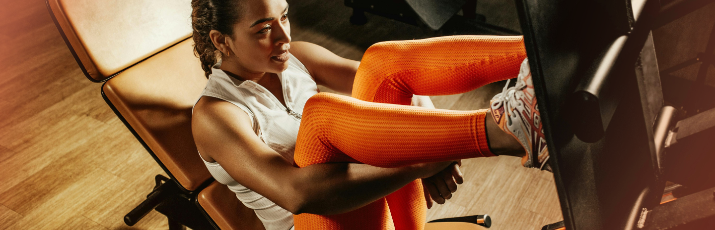 Woman in orange leggings and white top exercises on a leg press machine at the gym. She looks focused and determined, with warm lighting and wood flooring.