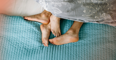 A man’s and a woman’s bare feet are visible on a bed during intimacy, partially covered by a blue quilt and a patterned grey blanket.